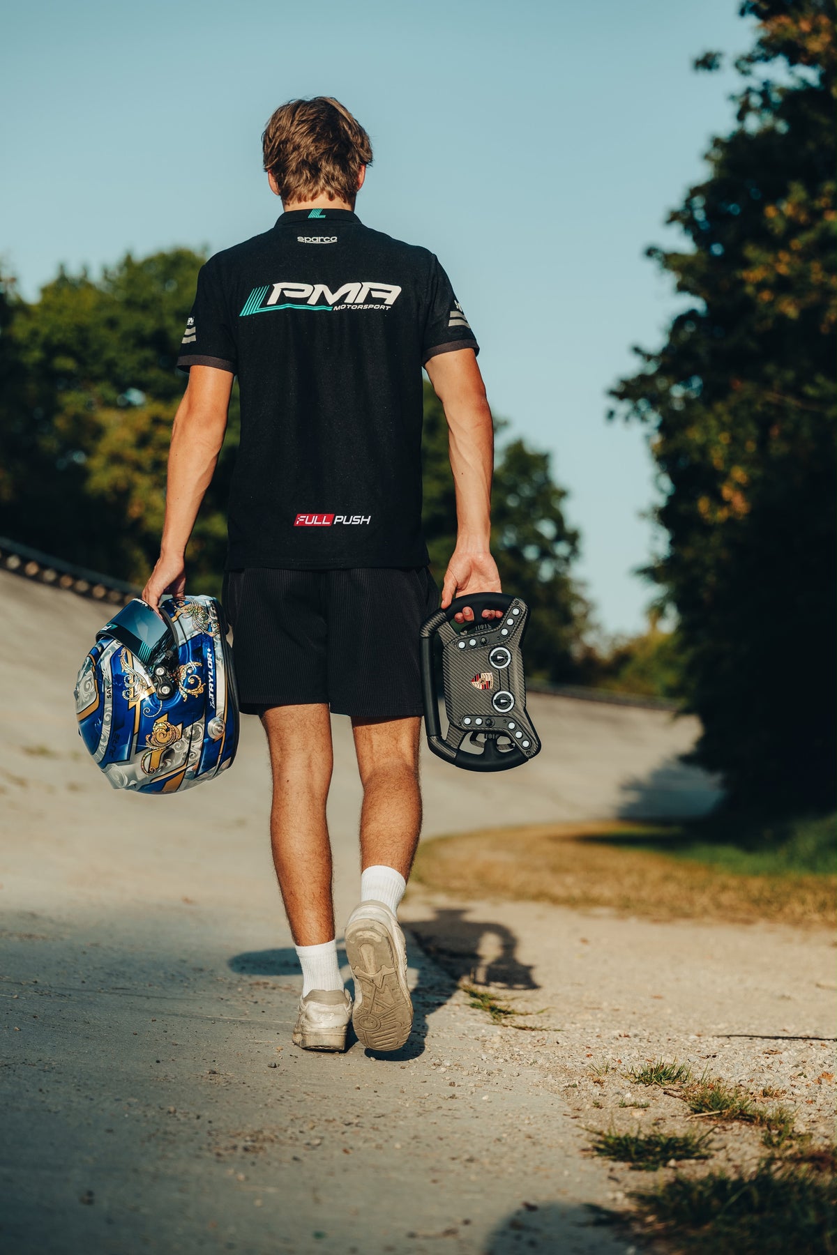 Race driver in a PMA Motorsport shirt with the Full Push logo walking down a banked track, carrying his helmet and a sim racing steering wheel